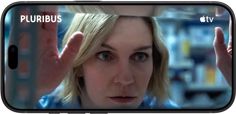 iPhone 18 Pro, mode paysage, affichant un gros plan d’une femme regardant à travers une étagère. Nom de la série : Pluribus