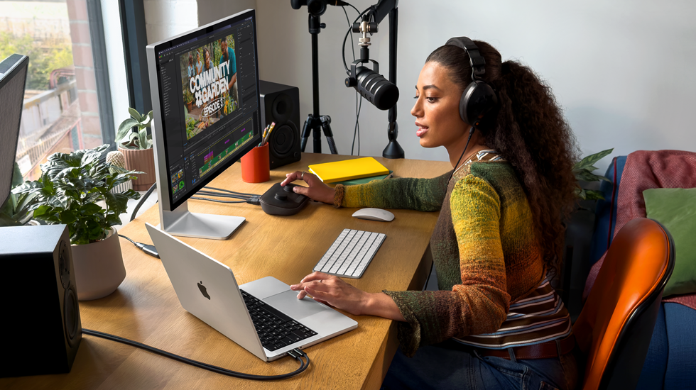 Une femme assise à son bureau chez elle travaille sur son MacBook Pro connecté à un écran externe, elle porte un casque et parle dans un micro