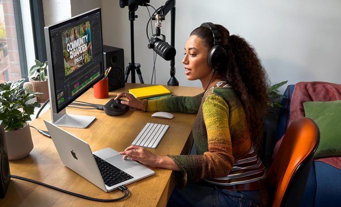Une femme assise à son bureau chez elle travaille sur son MacBook Pro connecté à un écran externe, elle porte un casque et parle dans un micro