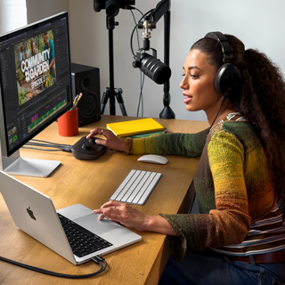 Une femme assise à son bureau chez elle travaille sur son MacBook Pro connecté à un écran externe, elle porte un casque et parle dans un micro
