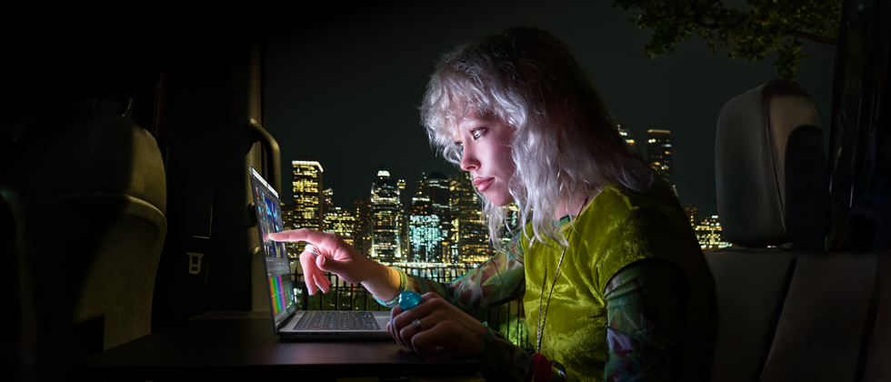 Une personne est assise sur la banquette arrière d’une camionnette, la porte passager ouverte laisse voir la ligne d’horizon d’une ville la nuit, un MacBook Pro avec Écran PROLED XDR est posé sur une table de travail, la personne, éclairée par l’écran, utilise la fonctionnalité tactile (Touch) pour monter une vidéo dans DaVinci Resolve Studio