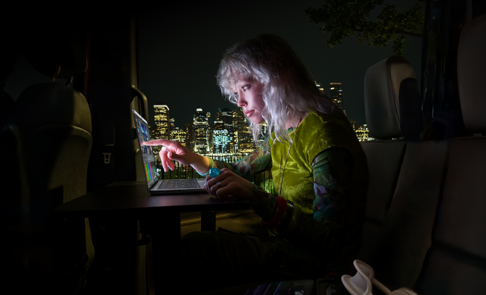 Une personne est assise sur la banquette arrière d’une camionnette, la porte passager ouverte laisse voir la ligne d’horizon d’une ville la nuit, un MacBook Pro avec Écran PROLED XDR est posé sur une table de travail, la personne, éclairée par l’écran, utilise la fonctionnalité tactile (Touch) pour monter une vidéo dans DaVinci Resolve Studio