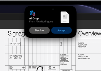 L’écran affiche la Dynamic Island avec une demande AirDrop dans une petite fenêtre au-dessus d’un fichier Adobe Acrobat ouvert